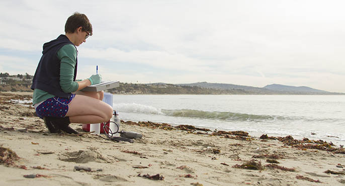 A field researcher surrounded by microbial contamination testing instruments squats on the sand at Doheny State Beach while taking notes.